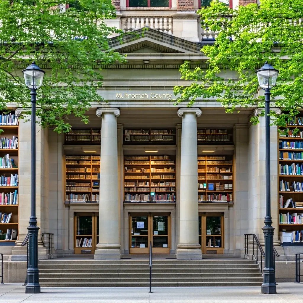 Front entrance of the Multnomah County Library with tall stone columns, shelves of books visible through the large windows, and leafy trees framing the historic building.