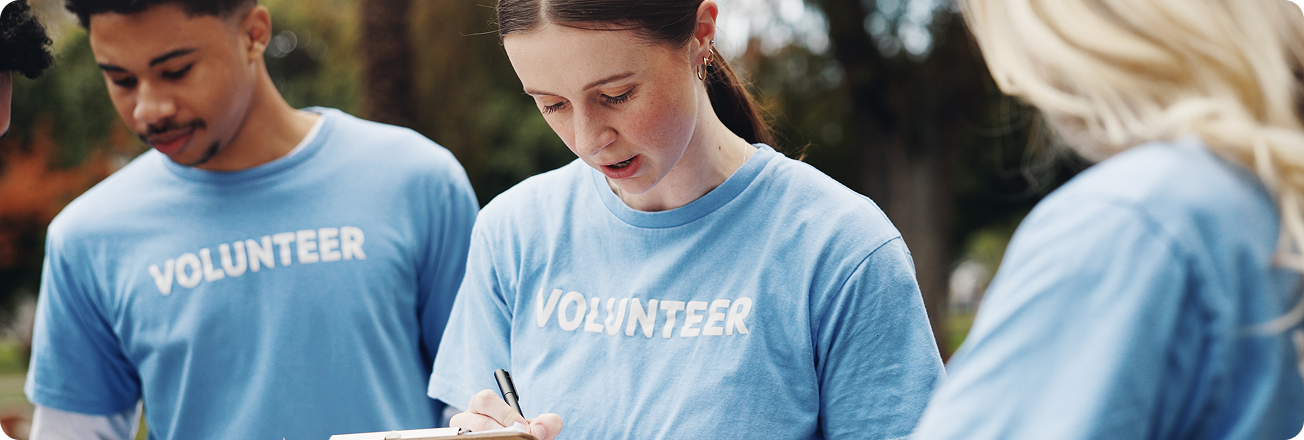Three volunteers in matching blue shirts collaborate outdoors, with one writing on a clipboard.