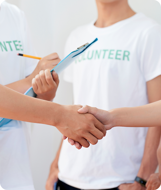 Two people shaking hands while others in volunteer T-shirts stand nearby, one holding a clipboard and pencil.