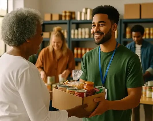 A young male volunteer at a food bank receives a box of food donations from an older woman, with shelves of supplies and other volunteers visible in the background.