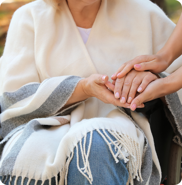Close-up of a caregiver gently holding the hands of an elderly woman wrapped in a blanket, symbolizing compassion and hospice care.