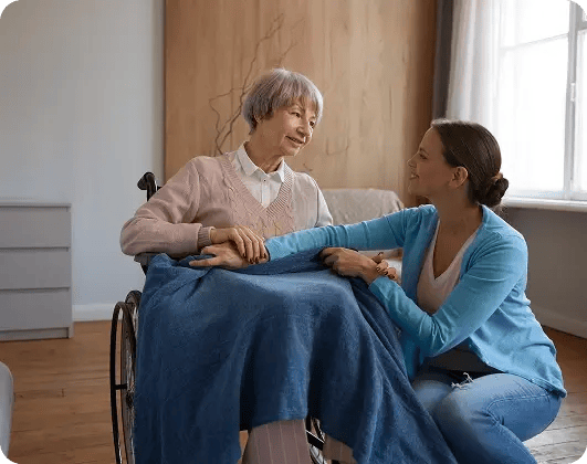 Volunteer holding hands and comforting an elderly woman in a wheelchair with a warm blanket in a hospice care setting.