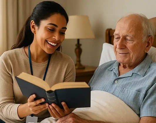 Young hospice volunteer in casual clothing reading a book to an elderly patient in bed, creating a comforting and supportive environment.