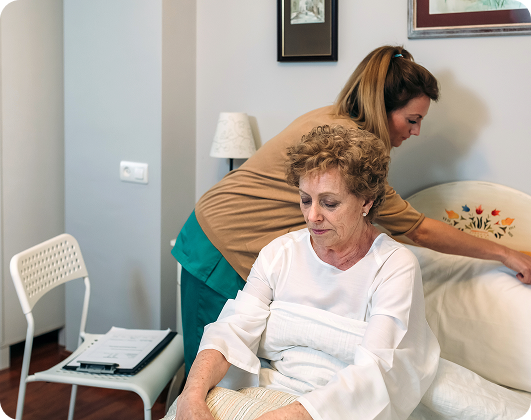 A caregiver adjusts bedding for an elderly woman sitting on the edge of a bed in a hospice care setting.