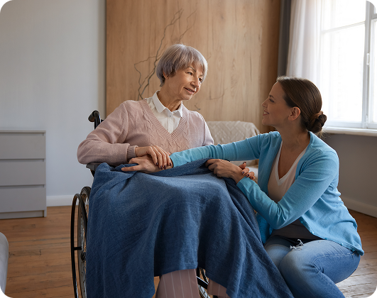 Volunteer holding hands and comforting an elderly woman in a wheelchair with a warm blanket in a hospice care setting.