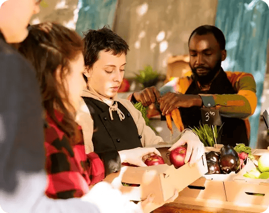 Volunteers packing apples, carrots, and fresh vegetables into food boxes at a community distribution table.