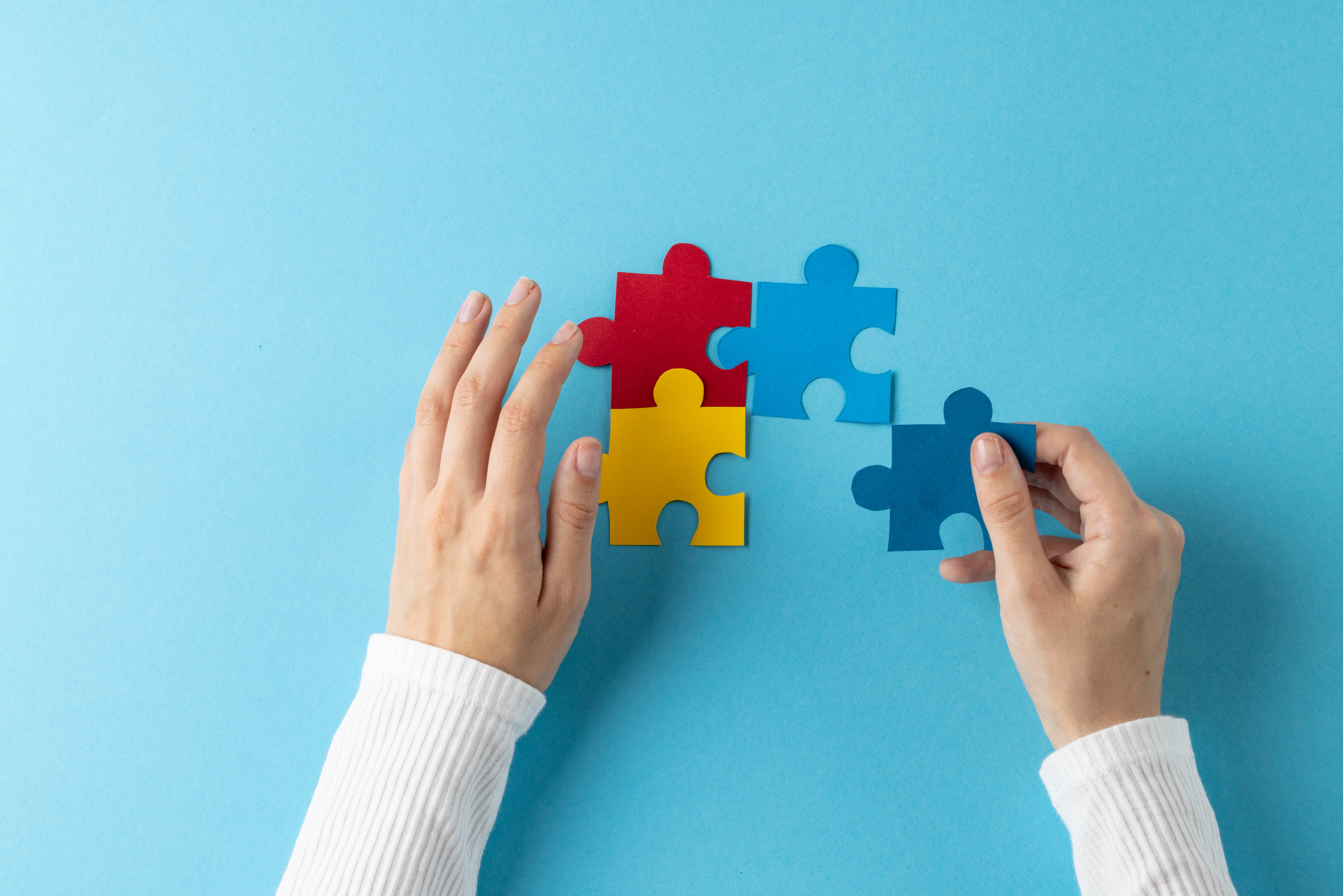 Hands connecting colourful puzzle pieces on a blue background.