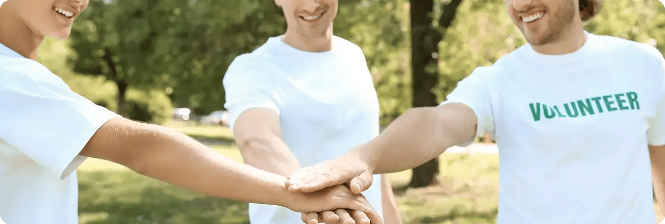 Three volunteers in matching white T-shirts stack hands in a show of teamwork outdoors.