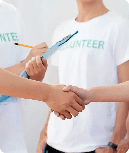 Two people shaking hands while others in volunteer T-shirts stand nearby, one holding a clipboard and pencil.