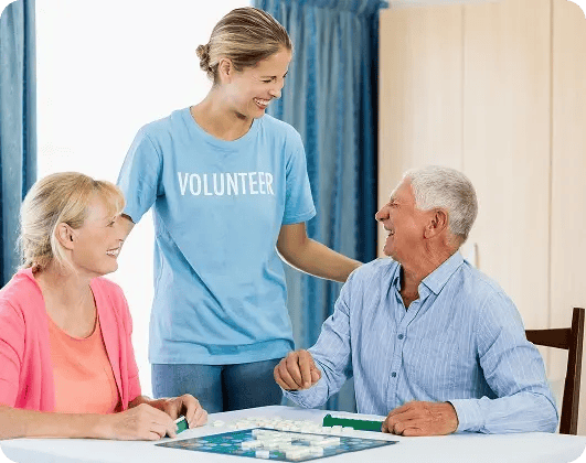 Smiling volunteer supporting two elderly people as they enjoy a board game together in a hospice care setting.