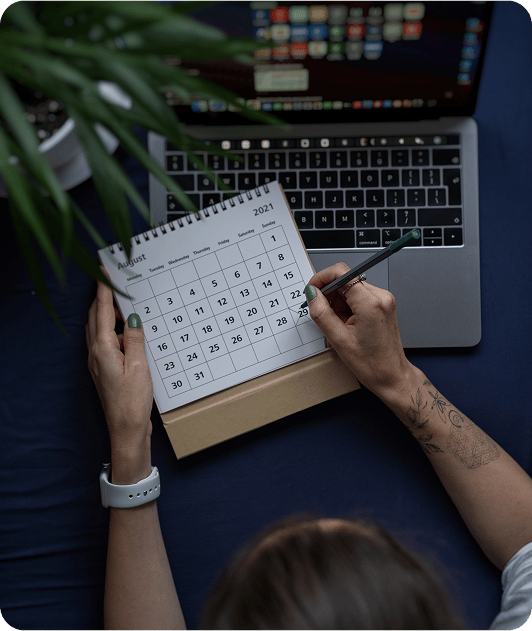 Person writing on a paper calendar while sitting in front of a laptop, representing volunteer scheduling and planning.