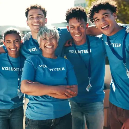 Diverse group of smiling volunteers wearing blue shirts with ID badges, standing arm-in-arm, representing community, collaboration, and impact through volunteerism.